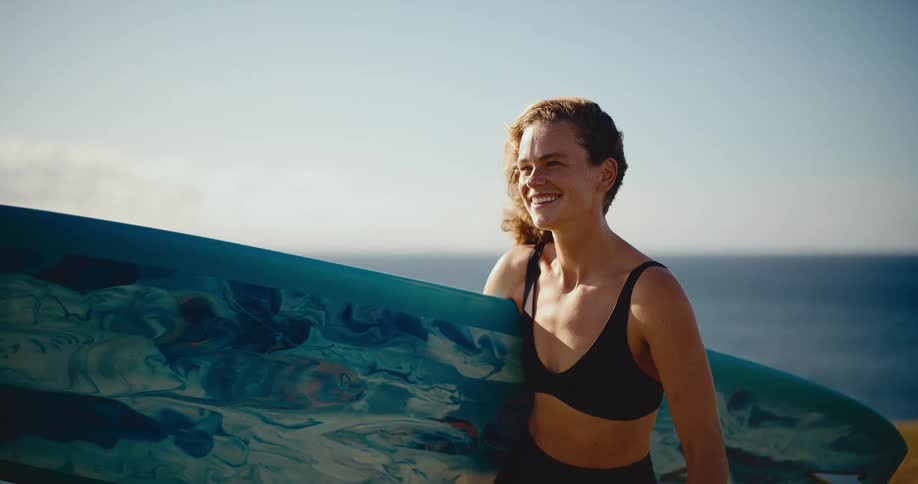 Happy young surfer woman smiling and holding her surfboard with the ocean in the background in slow motion