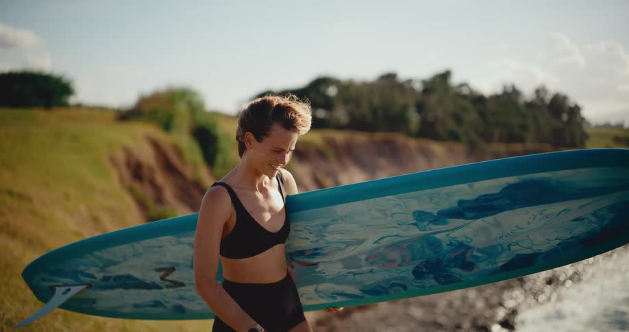 Cinematic slow motion of a smiling young woman with short hair carrying her longboard surfboard along a grassy coastline at golden hour