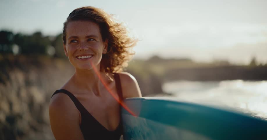Close up of a smiling female surfer holding her board as her hair blows in the wind at sunset in slow motion