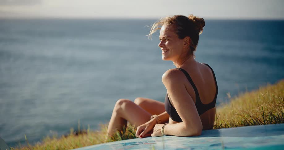 A beautiful surfer girl sits with her board on a grassy cliff, smiling as she watches the ocean at sunset