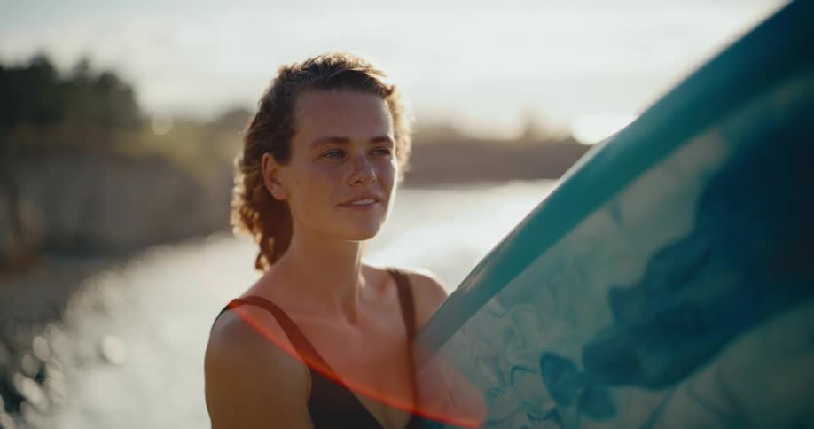 Slow motion close-up portrait of a happy young surfer woman smiling and holding her surfboard at the beach with the ocean in the background at sunset