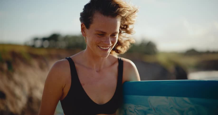 Portrait of a beautiful smiling surfer girl holding her surfboard with the wind in her hair at sunset in slow motion