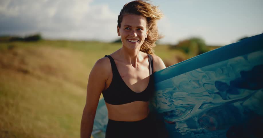 Slow motion portrait of a beautiful smiling woman holding her surfboard on a sunny day with her hair blowing in the wind
