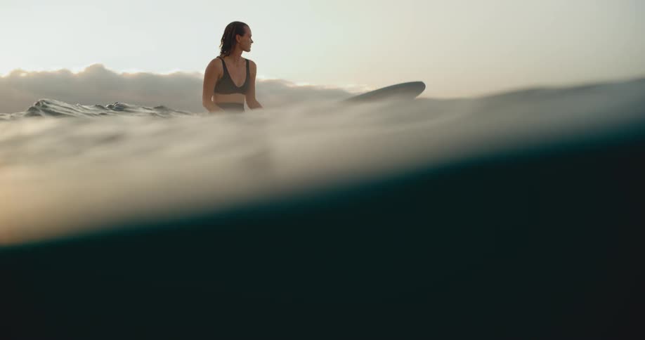 Beautiful smiling woman sitting on her surfboard in the ocean at sunset, waiting for a wave in slow motion