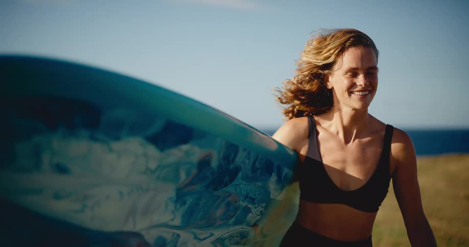 Beautiful smiling surfer girl walks with her surfboard along a grassy coastline at sunset in slow motion