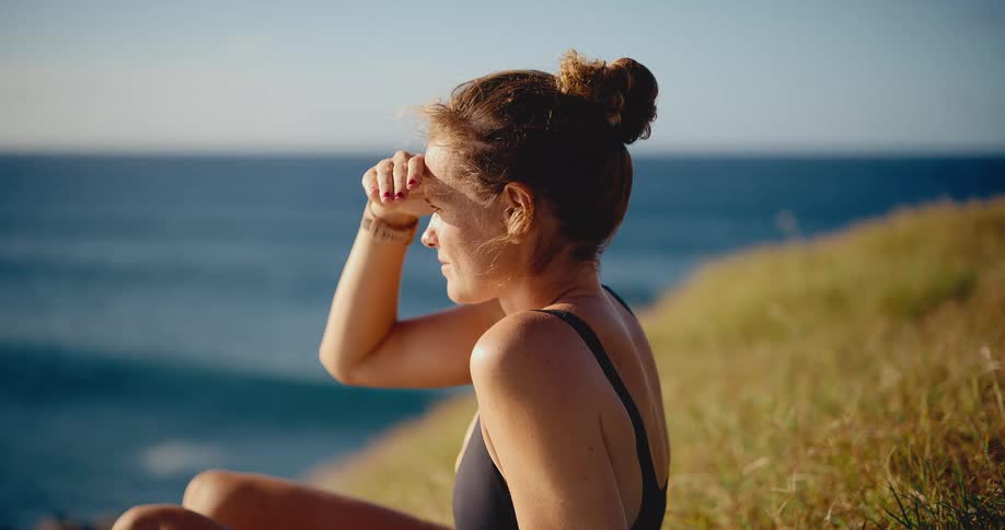 A serene woman in a black swimsuit shields her eyes from the sun as she sits on a grassy cliff watching the peaceful ocean
