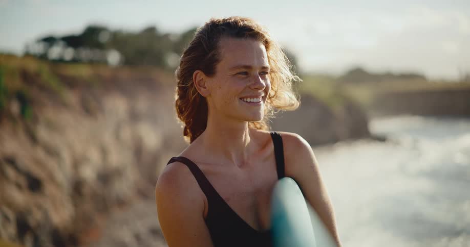 Portrait of a beautiful smiling woman holding her surfboard and looking out at the ocean at sunset