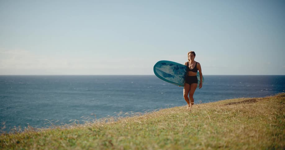 A smiling female surfer in a bikini walks up a grassy hill carrying her surfboard with the vast blue ocean in the background on a sunny day