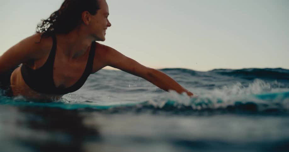A happy woman paddles on her surfboard in the ocean, smiling as she enjoys a beautiful sunset session in slow motion