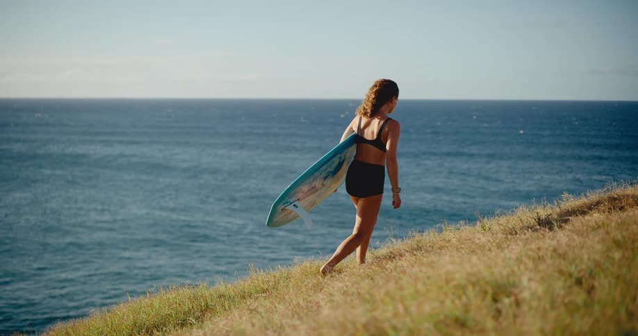 Barefoot surfer girl walks up a golden grassy hill with her surfboard, looking out at the endless blue ocean in slow motion
