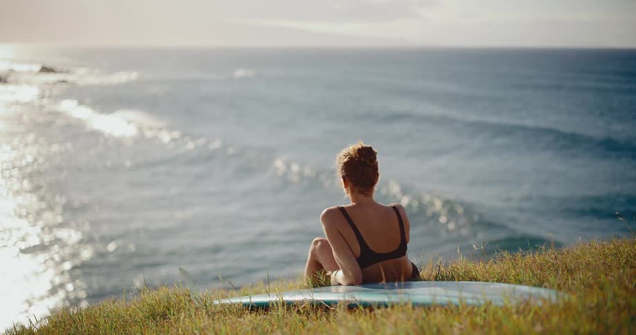 A beautiful surfer girl sits peacefully on a grassy cliff with her surfboard, watching the ocean waves glisten at sunset