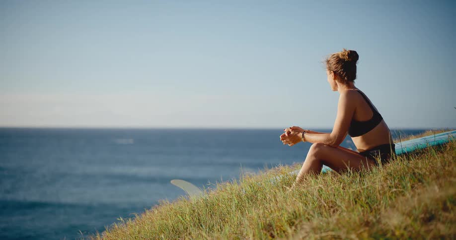 Beautiful young surfer woman sits peacefully on a grassy cliffside overlooking the ocean at golden hour