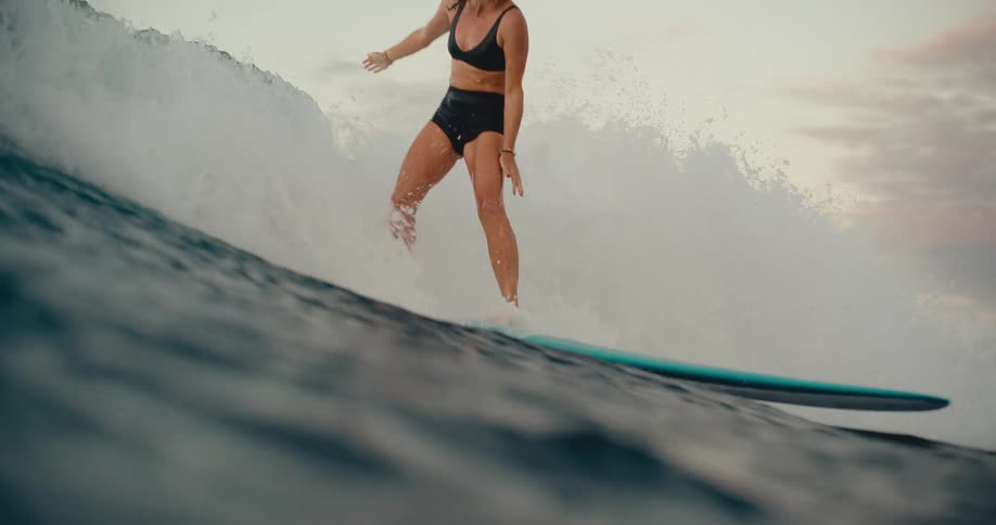 A smiling woman gracefully rides a large, powerful ocean wave on her longboard at dusk in slow motion