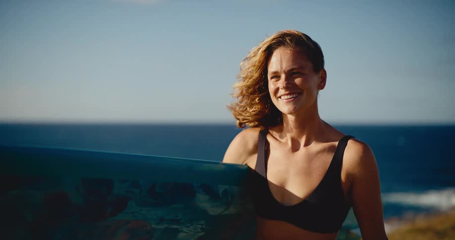A beautiful young woman surfer smiles while holding her board on a grassy cliff, with her hair blowing in the wind against an ocean backdrop