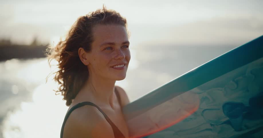 A beautiful female surfer with freckles smiles serenely while holding her surfboard on the beach during a windy golden hour sunset