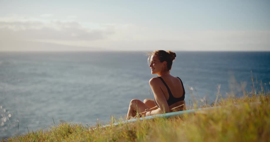 A beautiful smiling surfer woman sits on her surfboard on a grassy cliff, watching the ocean at sunset
