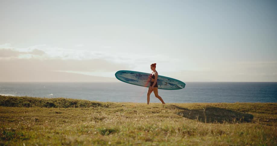 A young woman carries her surfboard along a grassy cliffside overlooking the ocean in beautiful golden light