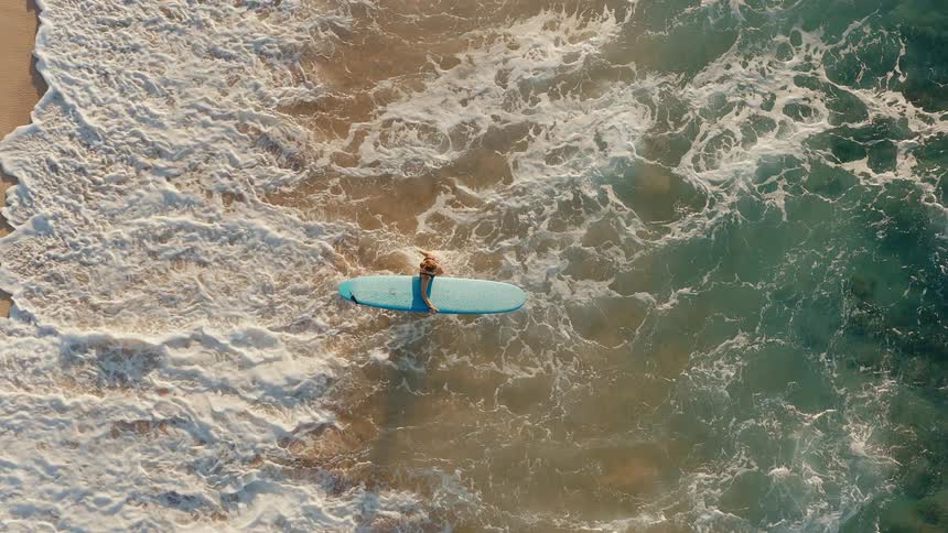 Top-down aerial slow-motion shot of a female surfer lying on her surfboard and paddling into the powerful ocean waves