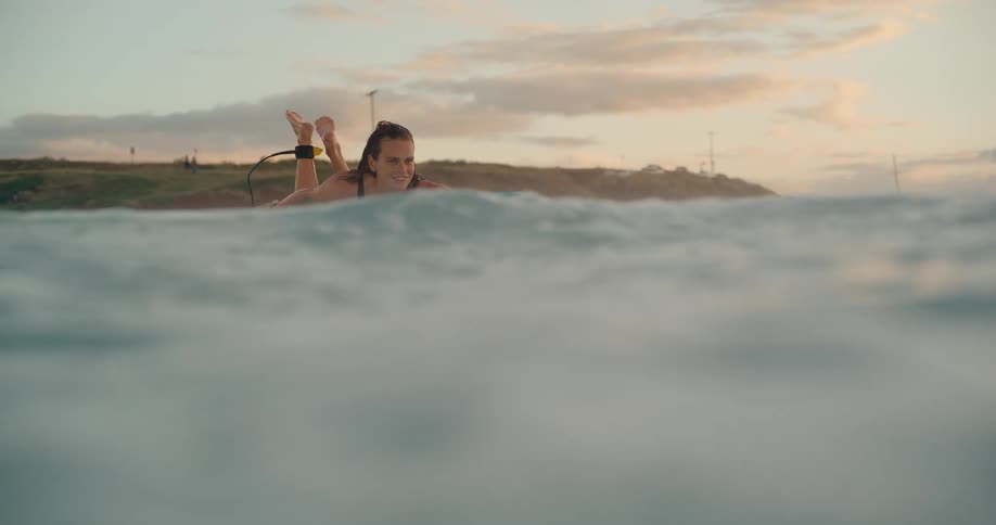 A happy young woman paddles her surfboard towards the camera at sunset, filmed in slow motion from a water-level perspective