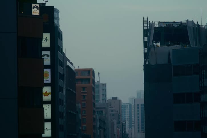 A static view down a quiet Tokyo street on an overcast day, with illuminated signs on one building and another under construction