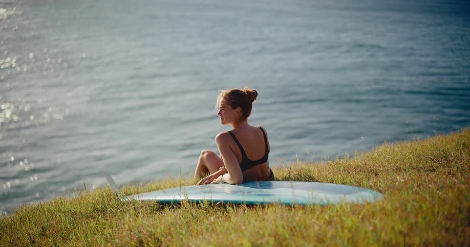 A female surfer in a bikini sits with her surfboard on a grassy hill, peacefully watching the ocean waves on a sunny day