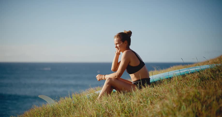 Beautiful surfer woman relaxing and smiling on a grassy hill with her surfboard overlooking the ocean at sunset