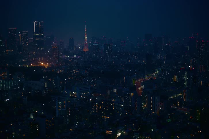 A wide, static aerial shot captures the sprawling Tokyo cityscape at night, with the iconic Tokyo Tower illuminated in the distance