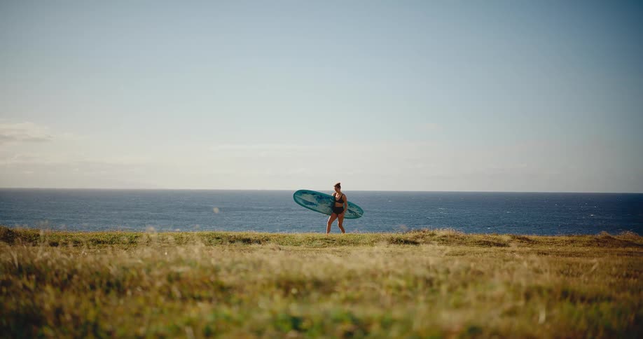 Female surfer with her surfboard walks along a grassy coastal bluff with the expansive blue ocean in the background