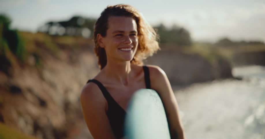 Close-up slow-motion shot of a beautiful young woman smiling while holding her surfboard on the beach during a golden sunset