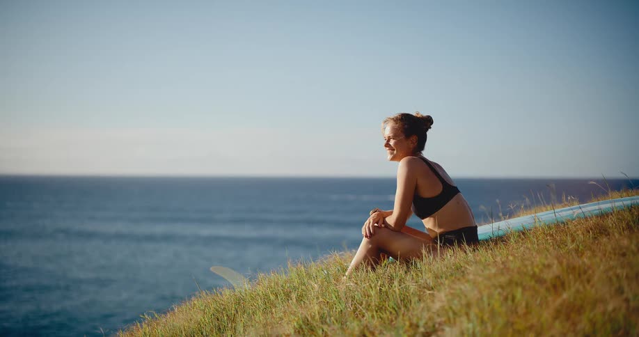 Happy young surfer woman sits on a grassy hill with her longboard, smiling as she watches the ocean waves on a sunny day