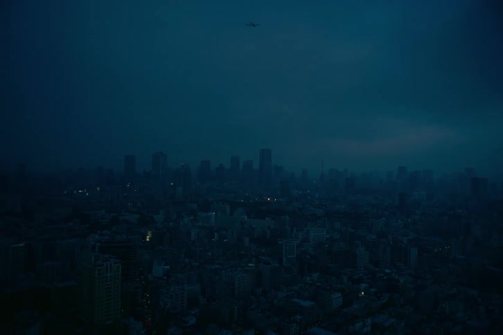 An airplane slowly crosses the dark blue sky above a sprawling, dimly lit cityscape just before dawn