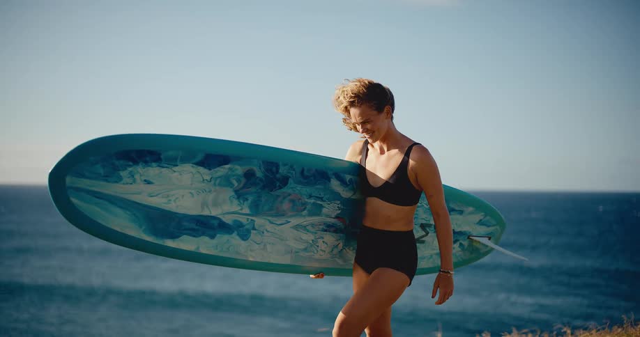 A smiling female surfer walks in slow motion with her longboard on a grassy cliff overlooking the ocean on a beautiful sunny day