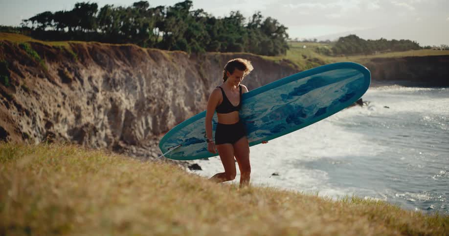 A happy female surfer walks up a windy, grassy cliffside with her longboard at sunset