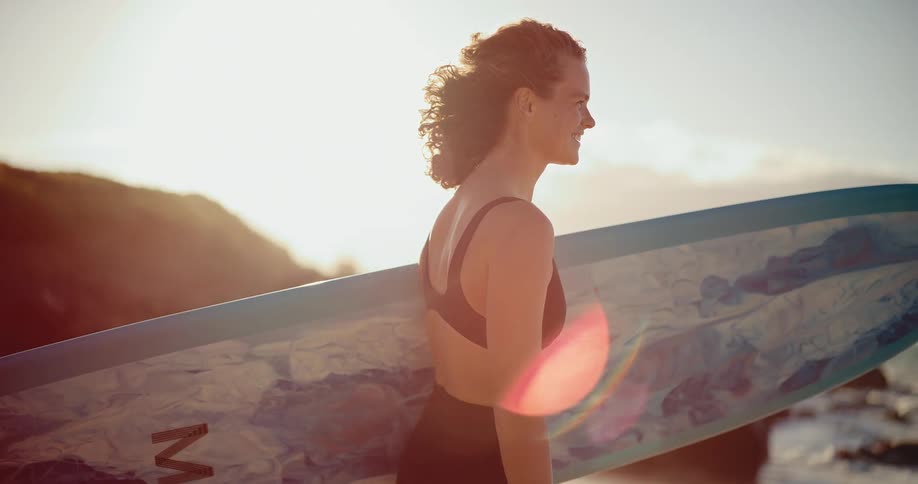 A happy woman holds her surfboard on the beach, watching the ocean waves at sunrise in slow motion with a beautiful lens flare