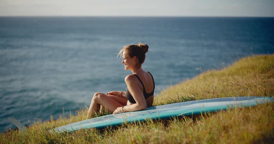 Happy attractive woman sitting on a grassy hill with her surfboard, watching the waves and enjoying a beautiful sunny day