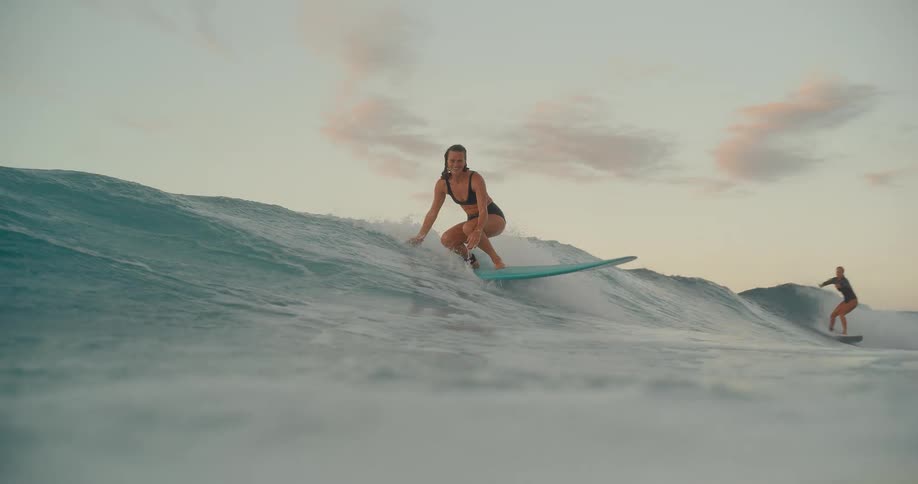 A smiling female surfer gracefully catches and rides a beautiful ocean wave at sunset, shot in stunning slow motion from the water
