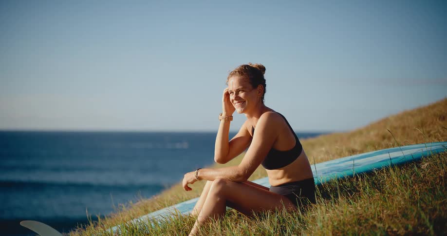 A beautiful young surfer woman relaxes and smiles on a grassy hill with her surfboard overlooking the ocean at sunset