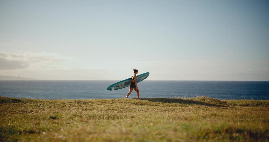A female surfer walks across a grassy clifftop with her longboard, heading towards the ocean on a beautiful sunny day