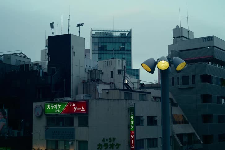 A gloomy, still shot of a Japanese city at dusk, featuring a yellow street lamp and neon signs
