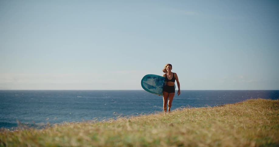 Woman carrying a surfboard walks along a sunny grassy coastal bluff overlooking the ocean