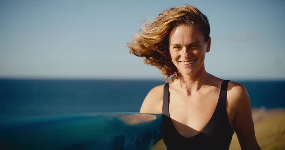 Portrait of a smiling young woman with her surfboard by the ocean, hair blowing in the wind in slow motion