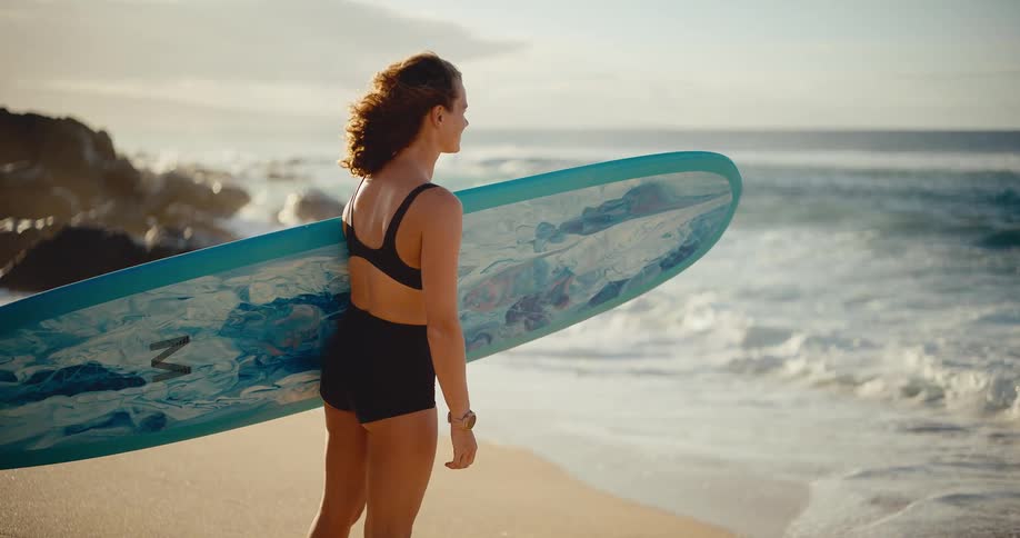 Female surfer holding a longboard stands on the beach watching the waves during a beautiful golden sunset