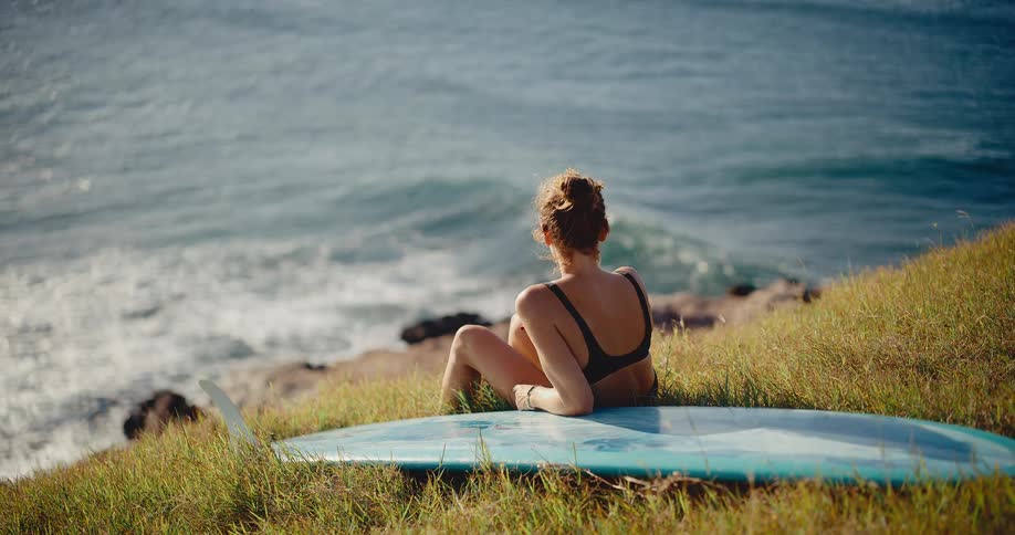 Smiling surfer girl relaxes on a grassy hill next to her surfboard and enjoys the view of the rolling ocean waves