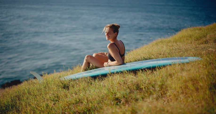 A beautiful surfer girl sits on her longboard on a grassy hill overlooking the ocean, enjoying the beautiful day and smiling