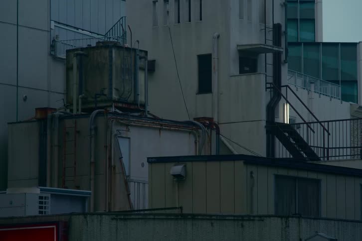 A static, moody shot of an industrial urban rooftop at dusk, showing weathered machinery and a single light glowing in the distance