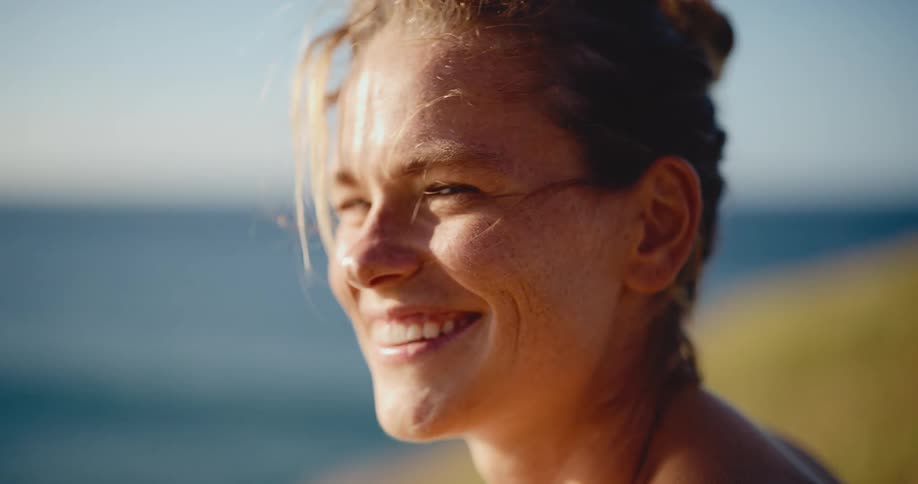 A serene portrait of a happy, freckled woman smiling as the golden hour sun shines on her face by the ocean
