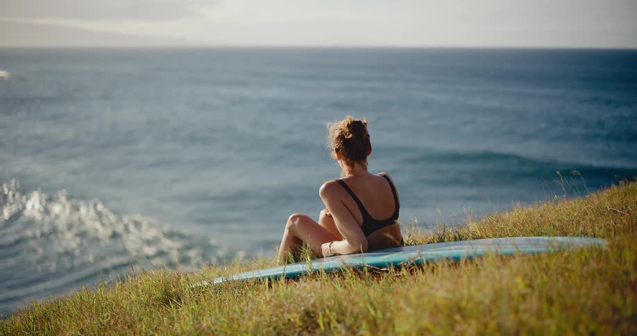 Happy young surfer woman sits on her surfboard on a grassy hill, smiling and watching the waves at sunset