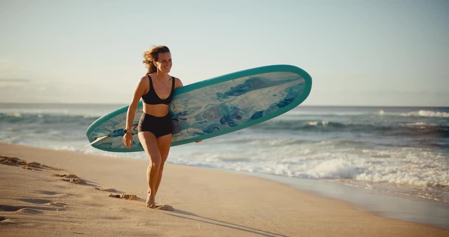 A happy and athletic woman smiles while carrying her surfboard and walking along a sandy beach at sunrise in slow motion