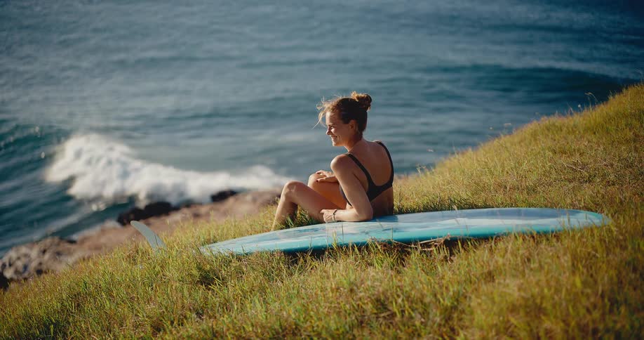 A beautiful surfer woman sits with her surfboard on a grassy hill, smiling as she watches the ocean waves