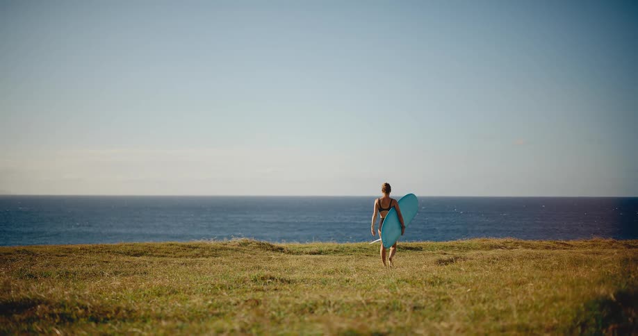 Female surfer carries her board through a grassy field towards the ocean on a sunny day